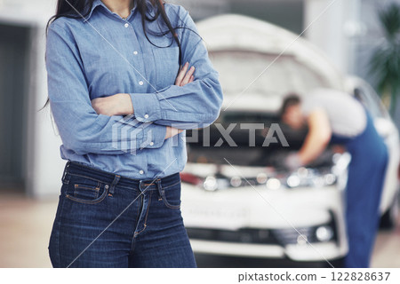 Woman at a car garage getting mechanical service. The mechanic works under the hood of the car 122828637