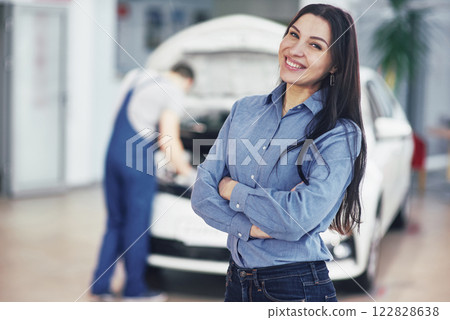 Woman at a car garage getting mechanical service. The mechanic works under the hood of the car 122828638