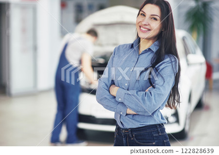 Woman at a car garage getting mechanical service. The mechanic works under the hood of the car Woman at a car garage getting mechanical service. The mechanic works under the hood of the car 122828639