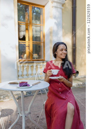 young African American woman in burgundy dress drinks tea in park in coffee shop 122829359