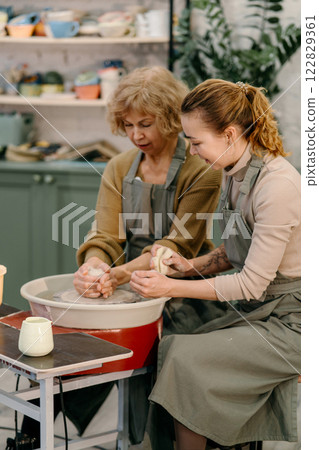 Hands-on learning, senior woman mastering pottery techniques using rotating wheel Mentor teaches pottery fundamentals, demonstrating sculpting skills on an electric wheel 122829361
