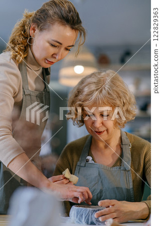 Teacher guides an elderly student, demonstrating sculpting techniques in a hands-on class. Senior woman practices clay modeling with help from a younger instructor in a ceramics studio. Teacher guides an elderly student, demonstrating sculpting techniques in a hands-on class. Senior woman practices clay modeling with help from a younger instructor in a ceramics studio. 122829363