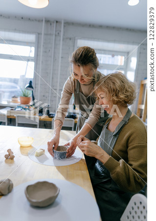 Happy retired student learns new skills, creating a handmade ceramic bowl. Female artist teaches an elderly woman how to shape clay at a hands-on master class Happy retired student learns new skills, creating a handmade ceramic bowl. Female artist teaches an elderly woman how to shape clay at a hands-on master class 122829379