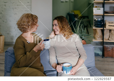 Mother and daughter, two Caucasian women of different ages, sit together on a couch with tea. The elderly mother and young daughter smile while enjoying a warm and relaxed conversation Mother and daughter, two Caucasian women of different ages, sit together on a couch with tea. The elderly mother and young daughter smile while enjoying a warm and relaxed conversation 122829387