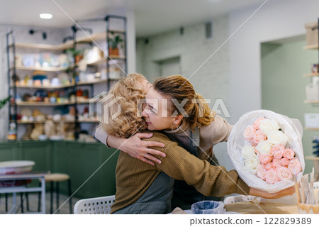 A young Caucasian woman surprises her elderly mother with a floral bouquet at a creative workshop. They celebrate a special event together, expressing love and joy in a cozy florist studio. A young Caucasian woman surprises her elderly mother with a floral bouquet at a creative workshop. They celebrate a special event together, expressing love and joy in a cozy florist studio. 122829389