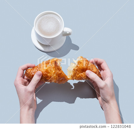 Hands breaking a fresh croissant over a blue background with a cup of cappuccino or latte coffee. 122831048