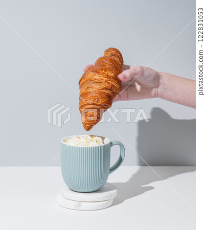 Hand dipping fresh croissant into cup of coffee with whipped cream on gray background with shadow. 122831053
