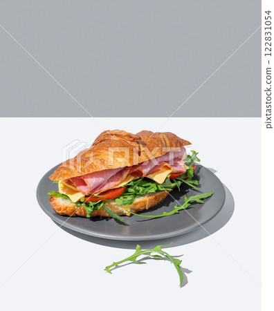 Croissant sandwich with ham, cheese and arugula on a dark plate on a light background with a shadow. Croissant sandwich with ham, cheese and arugula on a dark plate on a light background with a shadow. 122831054