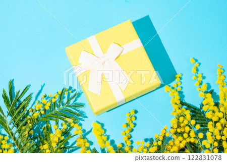 Yellow gift box on blue background with mimosa flowers and sunlight. Yellow gift box on blue background with mimosa flowers and sunlight. 122831078