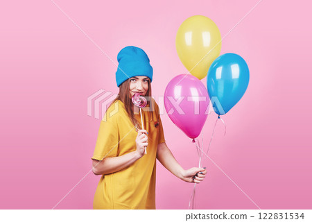 Cute funny girl in blue cap portrait holds an air colorful balloons and lollipop smiling on pink background. Beautiful multicultural Caucasian girl smiling happy Cute funny girl in blue cap portrait holds an air colorful balloons and lollipop smiling on pink background. Beautiful multicultural Caucasian girl smiling happy 122831534