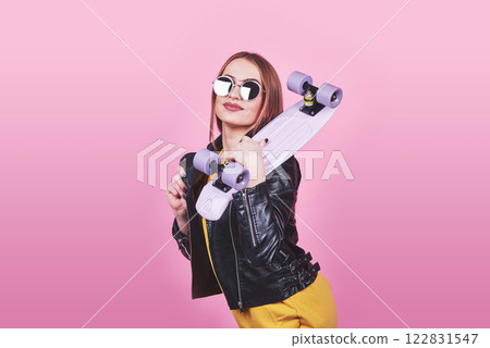 Attractive tanned girl with smile in face wears black leather jacket posing with pleasure. Indoor photo of lovely woman in sunglasses standing with skateboard on blue background Attractive tanned girl with smile in face wears black leather jacket posing with pleasure. Indoor photo of lovely woman in sunglasses standing with skateboard on blue background 122831547