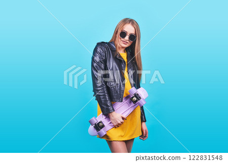 Attractive tanned girl with smile in face wears black leather jacket posing with pleasure. Indoor photo of lovely woman in sunglasses standing with skateboard on blue background 122831548