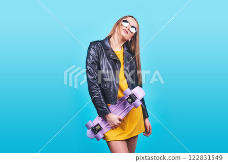 Attractive tanned girl with smile in face wears black leather jacket posing with pleasure. Indoor photo of lovely woman in sunglasses standing with skateboard on blue background Attractive tanned girl with smile in face wears black leather jacket posing with pleasure. Indoor photo of lovely woman in sunglasses standing with skateboard on blue background 122831549