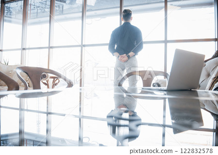 Young business executive and leader standing confidently, in the the top floor office, looking at the city below through large windows Young business executive and leader standing confidently, in the the top floor office, looking at the city below through large windows 122832578
