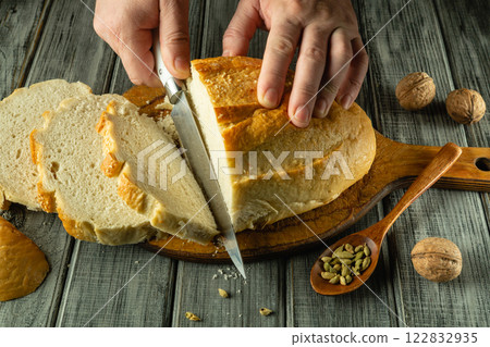 Hands expertly slice a golden-brown loaf of bread on a wooden board. Whole walnuts and spices rest nearby, adding a warm touch to this homely kitchen moment Hands expertly slice a golden-brown loaf of bread on a wooden board. Whole walnuts and spices rest nearby, adding a warm touch to this homely kitchen moment 122832935