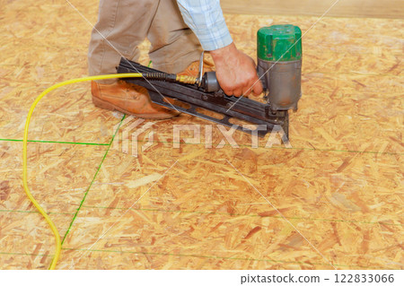 Construction worker operates nail pneumatic gun to secure flooring material, focusing on precision, safety. 122833066