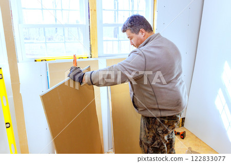 Carpenter cuts gypsum board, sheetrock sheets while renovating room filled with natural light. 122833077