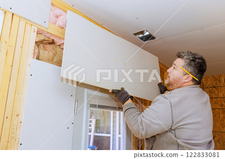 Worker carefully mounts drywall gypsum board on wooden framing in home interior. Worker carefully mounts drywall gypsum board on wooden framing in home interior. 122833081