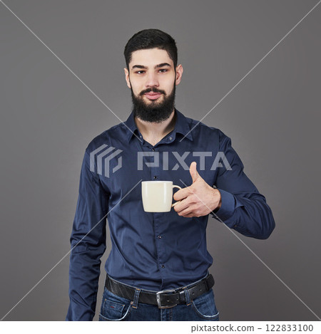 Handsome bearded man with stylish hair beard and mustache on serious face in shirt holding white cup or mug drinking tea or coffee in studio on grey background 122833100