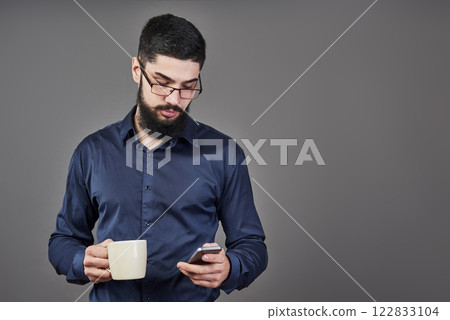 Handsome bearded man with stylish hair beard and mustache on serious face in shirt holding phone and white cup or mug drinking tea or coffee in studio on grey background Handsome bearded man with stylish hair beard and mustache on serious face in shirt holding phone and white cup or mug drinking tea or coffee in studio on grey background 122833104