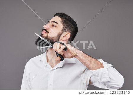 No more beard. Portrait of handsome young man cutting his beard with scissors and looking at camera while standing against grey background. New trend No more beard. Portrait of handsome young man cutting his beard with scissors and looking at camera while standing against grey background. New trend 122833140