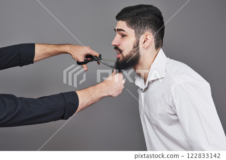 No more beard. Portrait of handsome young man cutting his beard with scissors and looking at camera while standing against grey background. New trend No more beard. Portrait of handsome young man cutting his beard with scissors and looking at camera while standing against grey background. New trend 122833142