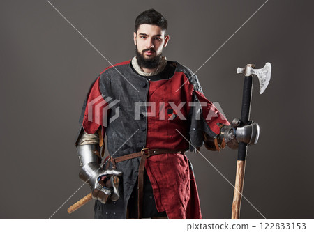 Medieval knight on grey background. Portrait of brutal dirty face warrior with chain mail armour red and black clothes and battle axe Medieval knight on grey background. Portrait of brutal dirty face warrior with chain mail armour red and black clothes and battle axe 122833153