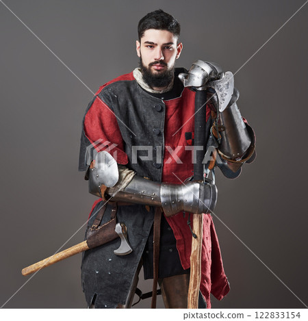 Medieval knight on grey background. Portrait of brutal dirty face warrior with chain mail armour red and black clothes and battle axe 122833154