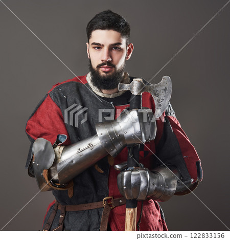Medieval knight on grey background. Portrait of brutal dirty face warrior with chain mail armour red and black clothes and battle axe 122833156