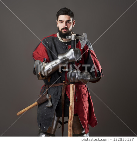 Medieval knight on grey background. Portrait of brutal dirty face warrior with chain mail armour red and black clothes and battle axe 122833157