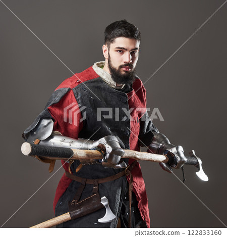 Medieval knight on grey background. Portrait of brutal dirty face warrior with chain mail armour red and black clothes and battle axe 122833160