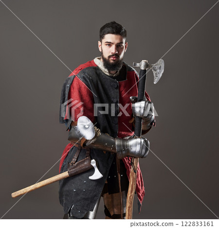 Medieval knight on grey background. Portrait of brutal dirty face warrior with chain mail armour red and black clothes and battle axe 122833161