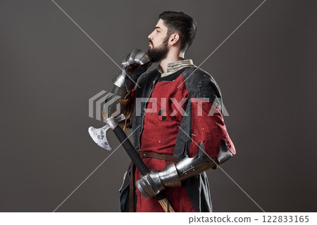 Medieval knight on grey background. Portrait of brutal dirty face warrior with chain mail armour red and black clothes and battle axe 122833165