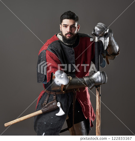 Medieval knight on grey background. Portrait of brutal dirty face warrior with chain mail armour red and black clothes and battle axe 122833167