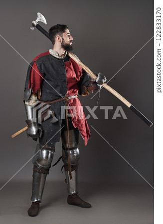 Medieval knight on grey background. Portrait of brutal dirty face warrior with chain mail armour red and black clothes and battle axe 122833170