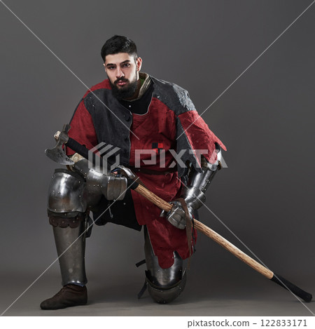 Medieval knight on grey background. Portrait of brutal dirty face warrior with chain mail armour red and black clothes and battle axe Medieval knight on grey background. Portrait of brutal dirty face warrior with chain mail armour red and black clothes and battle axe 122833171