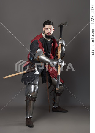 Medieval knight on grey background. Portrait of brutal dirty face warrior with chain mail armour red and black clothes and battle axe 122833172