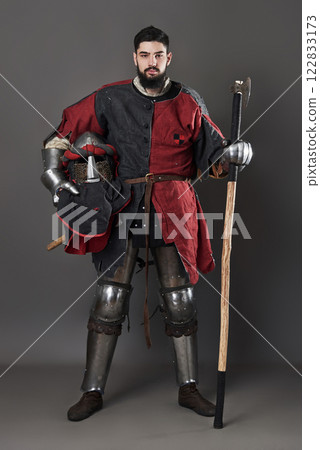 Medieval knight on grey background. Portrait of brutal dirty face warrior with chain mail armour red and black clothes and battle axe 122833173
