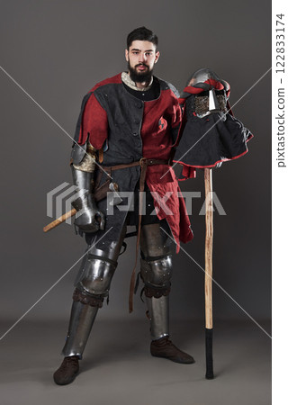 Medieval knight on grey background. Portrait of brutal dirty face warrior with chain mail armour red and black clothes and battle axe Medieval knight on grey background. Portrait of brutal dirty face warrior with chain mail armour red and black clothes and battle axe 122833174