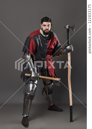 Medieval knight on grey background. Portrait of brutal dirty face warrior with chain mail armour red and black clothes and battle axe 122833175
