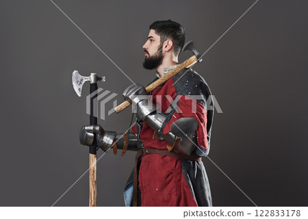 Medieval knight on grey background. Portrait of brutal dirty face warrior with chain mail armour red and black clothes and battle axe 122833178