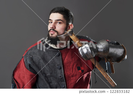 Medieval knight on grey background. Portrait of brutal dirty face warrior with chain mail armour red and black clothes and battle axe 122833182