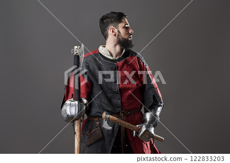 Medieval knight on grey background. Portrait of brutal dirty face warrior with chain mail armour red and black clothes and battle axe 122833203