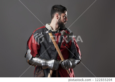 Medieval knight on grey background. Portrait of brutal dirty face warrior with chain mail armour red and black clothes and battle axe 122833204