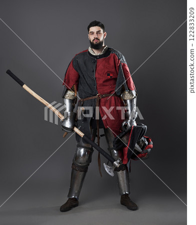 Medieval knight on grey background. Portrait of brutal dirty face warrior with chain mail armour red and black clothes and battle axe 122833209