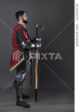 Medieval knight on grey background. Portrait of brutal dirty face warrior with chain mail armour red and black clothes and battle axe 122833218