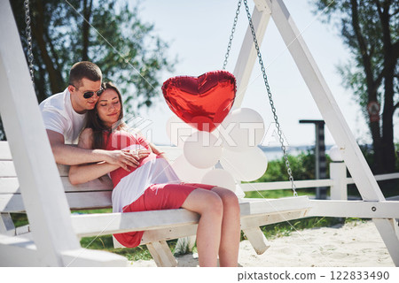 waiting baby. pregnant woman with beloved husband sit on a bench. Husband hand embraces a round belly wife. Parenthood. pregnant woman in a white dress in cherry with a red bow. close-up. nine months 122833490