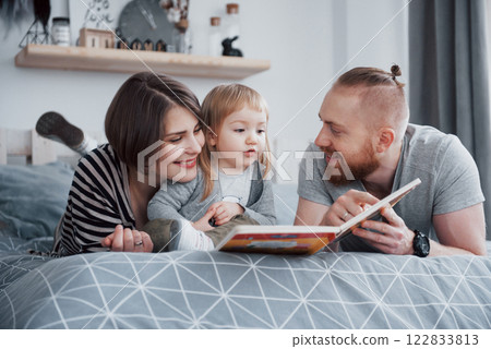 Father, Mother and Little Daughter Reading Children's Book on a Sofa in the Living Room. Happy big family read an interesting book on a festive day. Parents love their children Father, Mother and Little Daughter Reading Children's Book on a Sofa in the Living Room. Happy big family read an interesting book on a festive day. Parents love their children 122833813