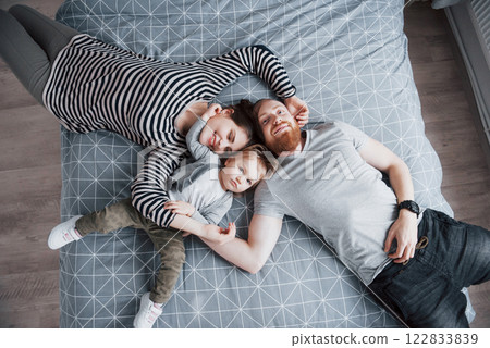 Top view of beautiful young mother, father and their daughter looking at camera and smiling while lying on bed head to head 122833839