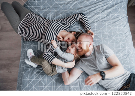 Top view of beautiful young mother, father and their daughter looking at camera and smiling while lying on bed head to head Top view of beautiful young mother, father and their daughter looking at camera and smiling while lying on bed head to head 122833842